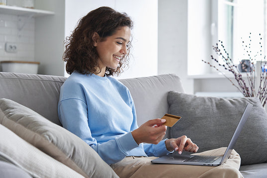 Woman sitting on a sofa using a laptop and holding a payment card beside text about every Apple purchase through TechForGood being carbon neutral.