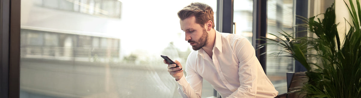 Man in white shirt using smartphone near office window with green plant